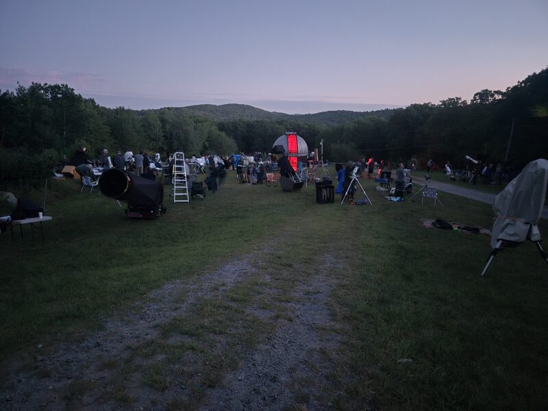 Observers wait with anticipation on an observing field bristling with telescopes as the Sun sets. A red glow can be seen coming from the small domed observatory at the center.