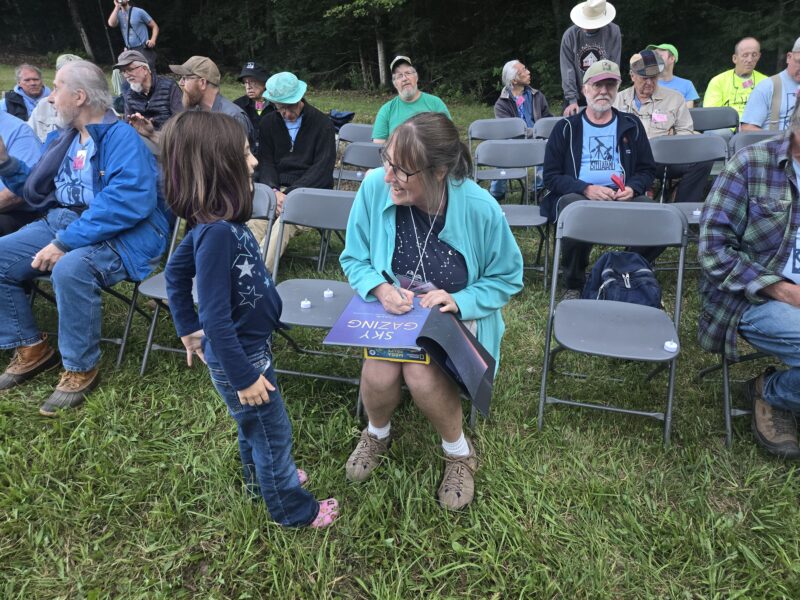 Meg Thacher sits on a plastic chair signing a copy of her book while the young girl who won the book in a raffle looks on.