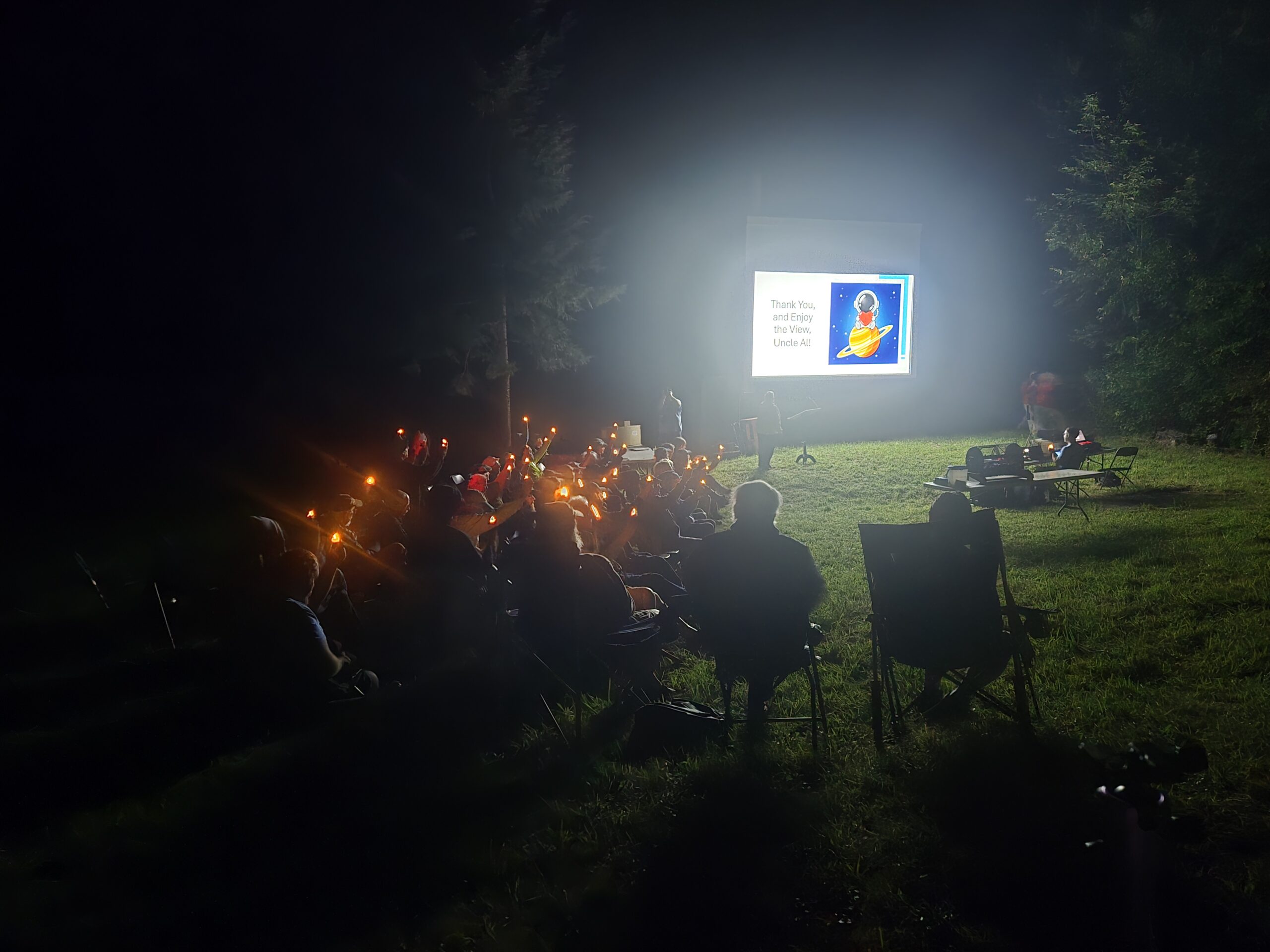 A group of Springfield Telescope Makers members sitting to the left of a white projection screen. The members are all holding glowing, battery-powered tea lights, and the projection screen has a picture of a cartoon astronaut sitting on Saturn holding a heart. The Screen reads 