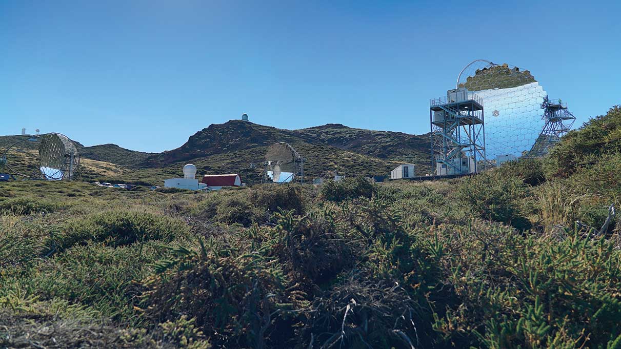 an outdoor shot of a grassy hill with a large circle with honeycomb shapes in it towering over other buildings and machinery