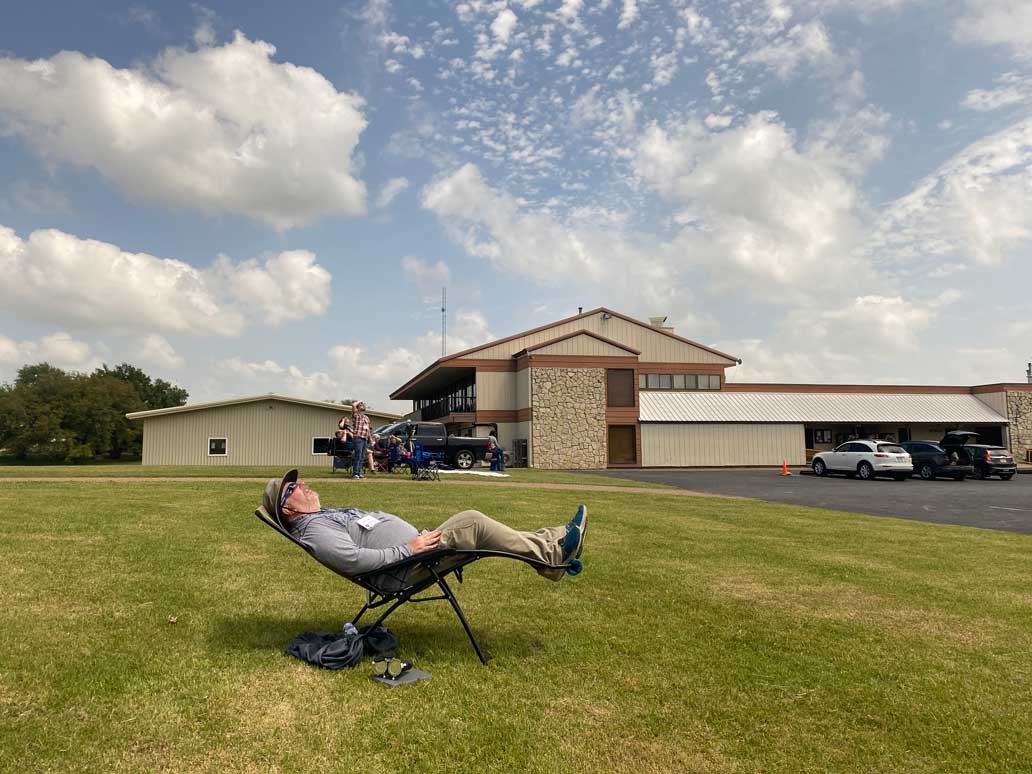 a man reclines in an outdoor chair wearing eclipse glasses in front of a building and a sky with clouds