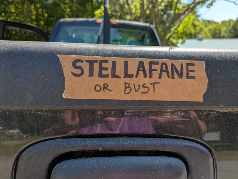 The tailgate of a black pickup truck with a strip of paper tape stuck to it with "Stellafane or Bust" written on it in black sharpie. 