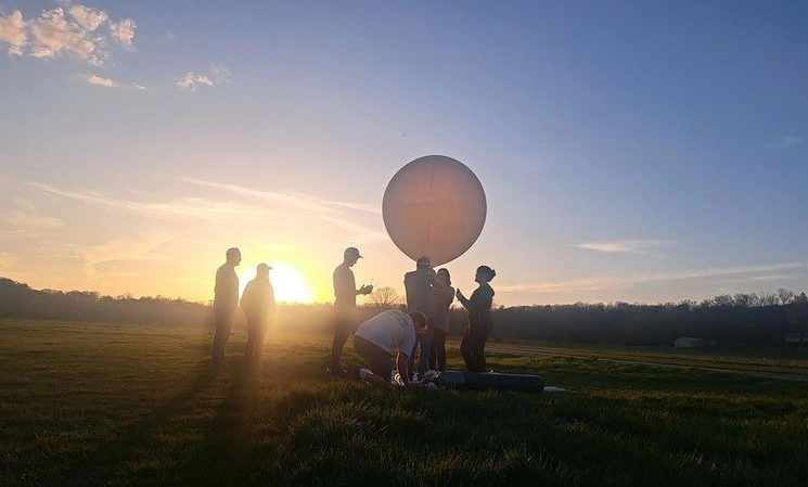 Students stand around white balloon with the Sun in the background