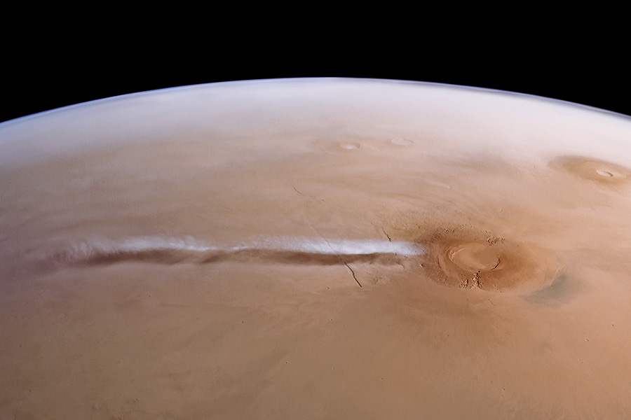Tan-coloured horizontal rectangle showing part of the surface of Mars imaged by an orbiting satellite. Above the curved horizon is space. To the lower right of the image is a volcano. Stretching to the left of the volcano is a long wisp of white cloud, which seems to emanate from the volcano. We also see the shadow of the cloud just below the cloud itself