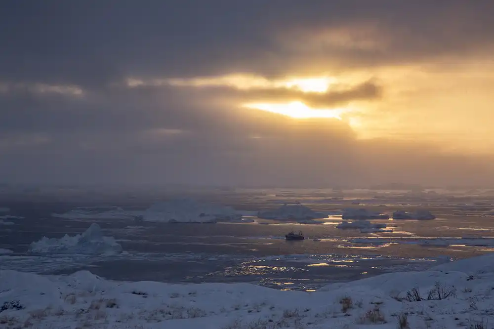 Golden sunlight pierces clouds over a bay filled with floating icebergs, and a ship for scale